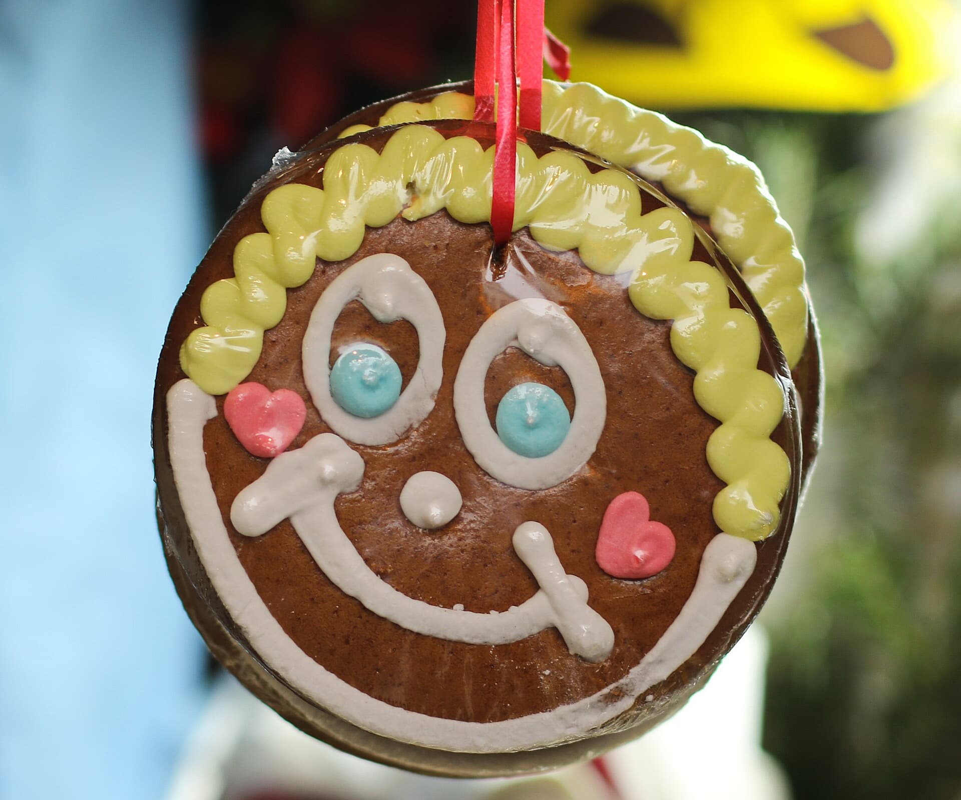 Runder Lebkuchen mit lachendem Gesicht aus weißem, gelbem, blauem und rosa Zuckerguss hängt an einem roten Band